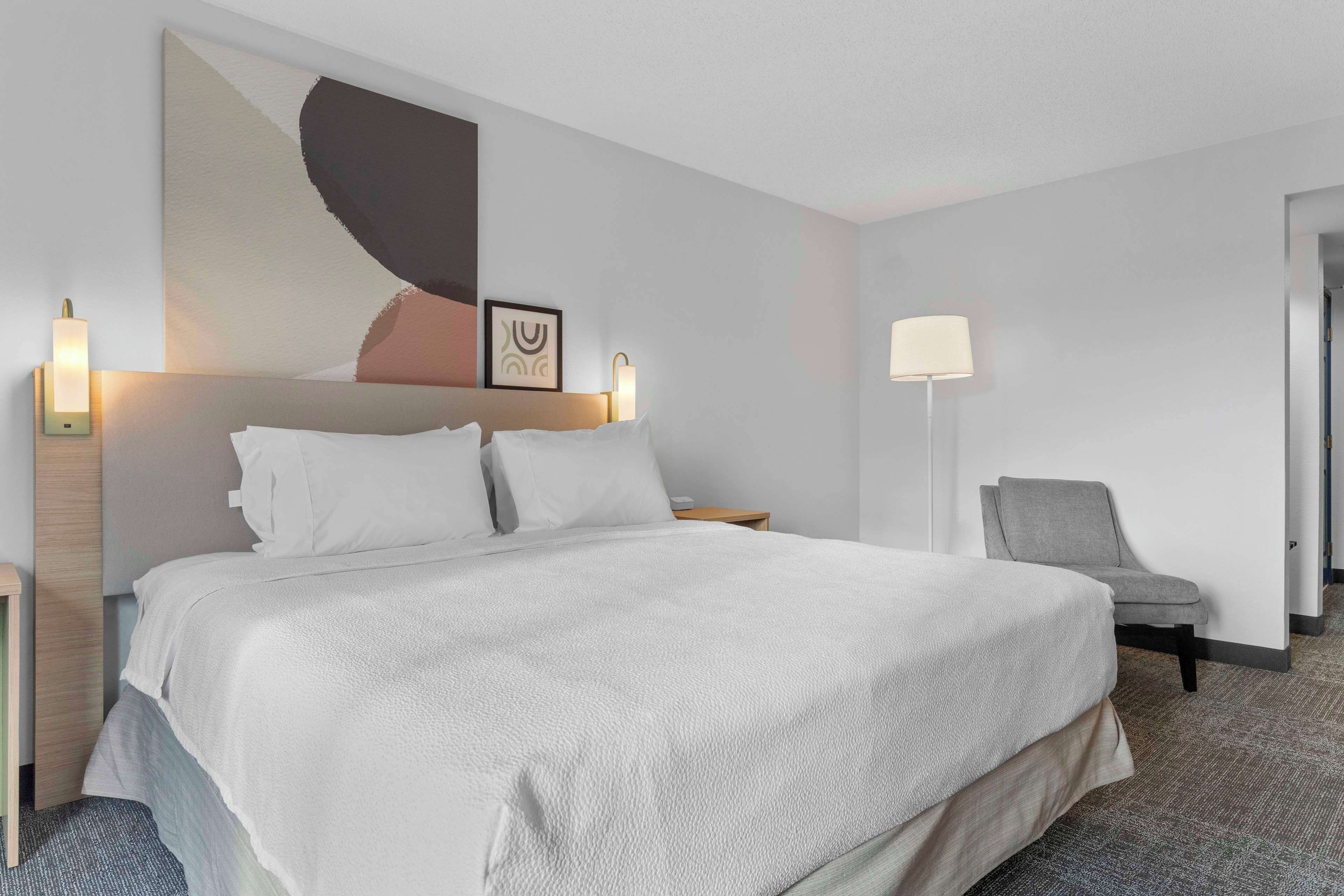 A hotel room with a large bed, two bedside lamps, a chair, and a wall art piece.