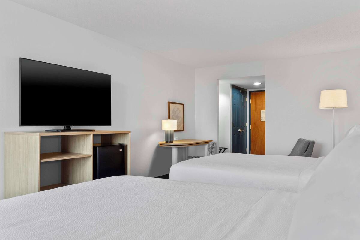 Une chambre d’hôtel avec deux lits, un téléviseur à écran plat, un petit réfrigérateur, un bureau avec une lampe et une porte donnant sur une autre chambre.