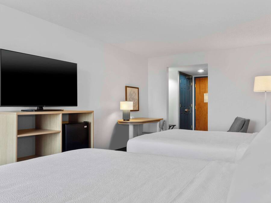 Ett hotellrum med två sängar, en platt-TV, ett litet kylskåp, ett skrivbord med en lampa och en dörr som leder till ett annat rum.