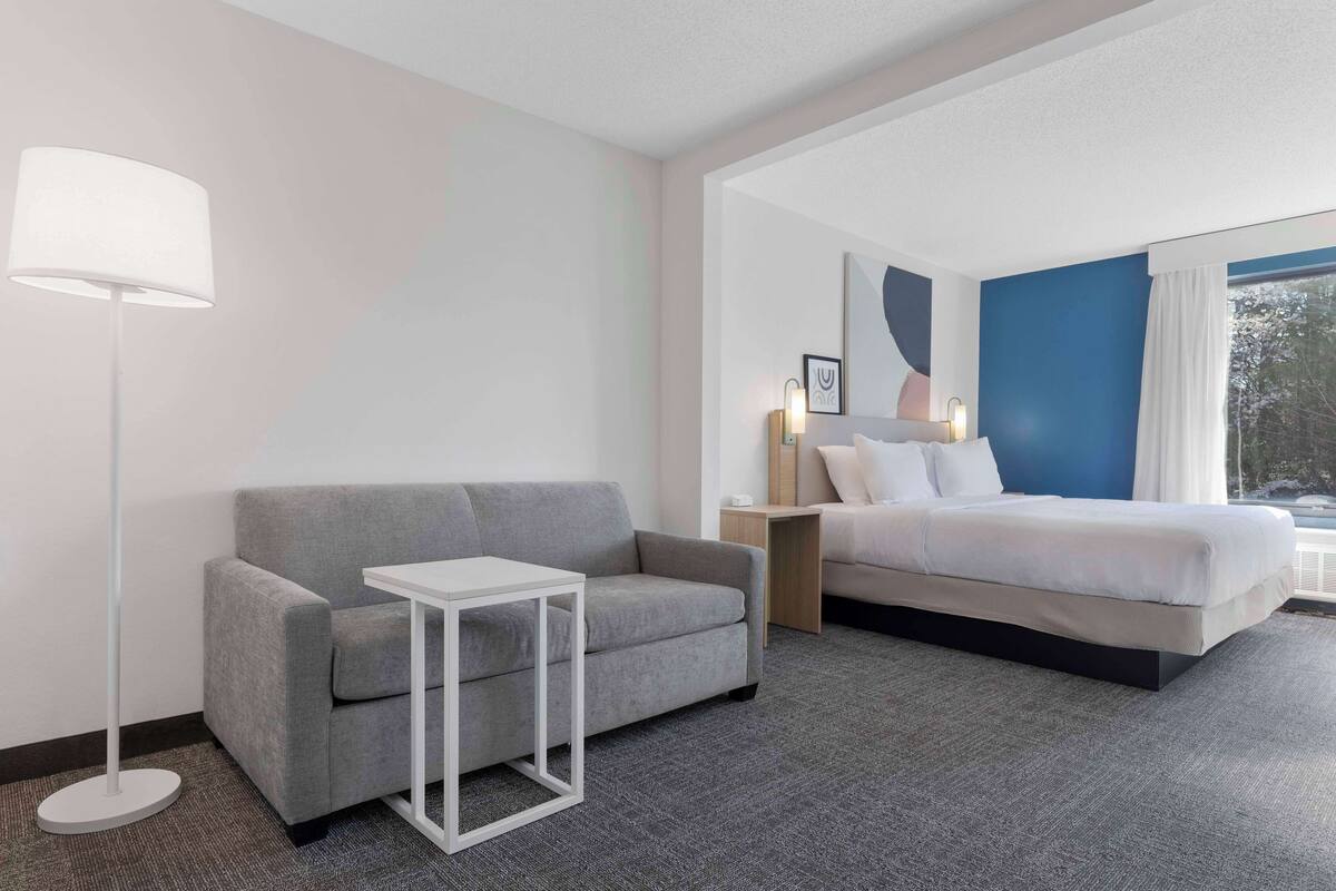 Une chambre d’hôtel moderne avec un canapé gris, une petite table blanche, un lit avec une literie blanche, une table de chevet avec une lampe et une grande fenêtre donnant sur des arbres.