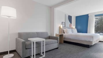 Una habitación de hotel moderna con un sofá gris, una pequeña mesa blanca, una cama con ropa de cama blanca, una mesita de noche con una lámpara y un ventanal con vistas a árboles.
