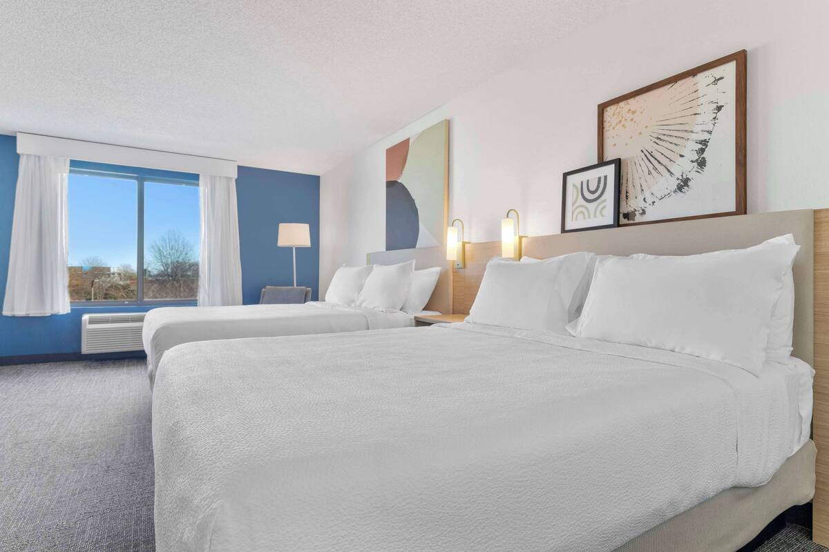 Une chambre d’hôtel avec deux lits, une grande fenêtre avec des rideaux, une lampe fixée au mur et des œuvres d’art.
