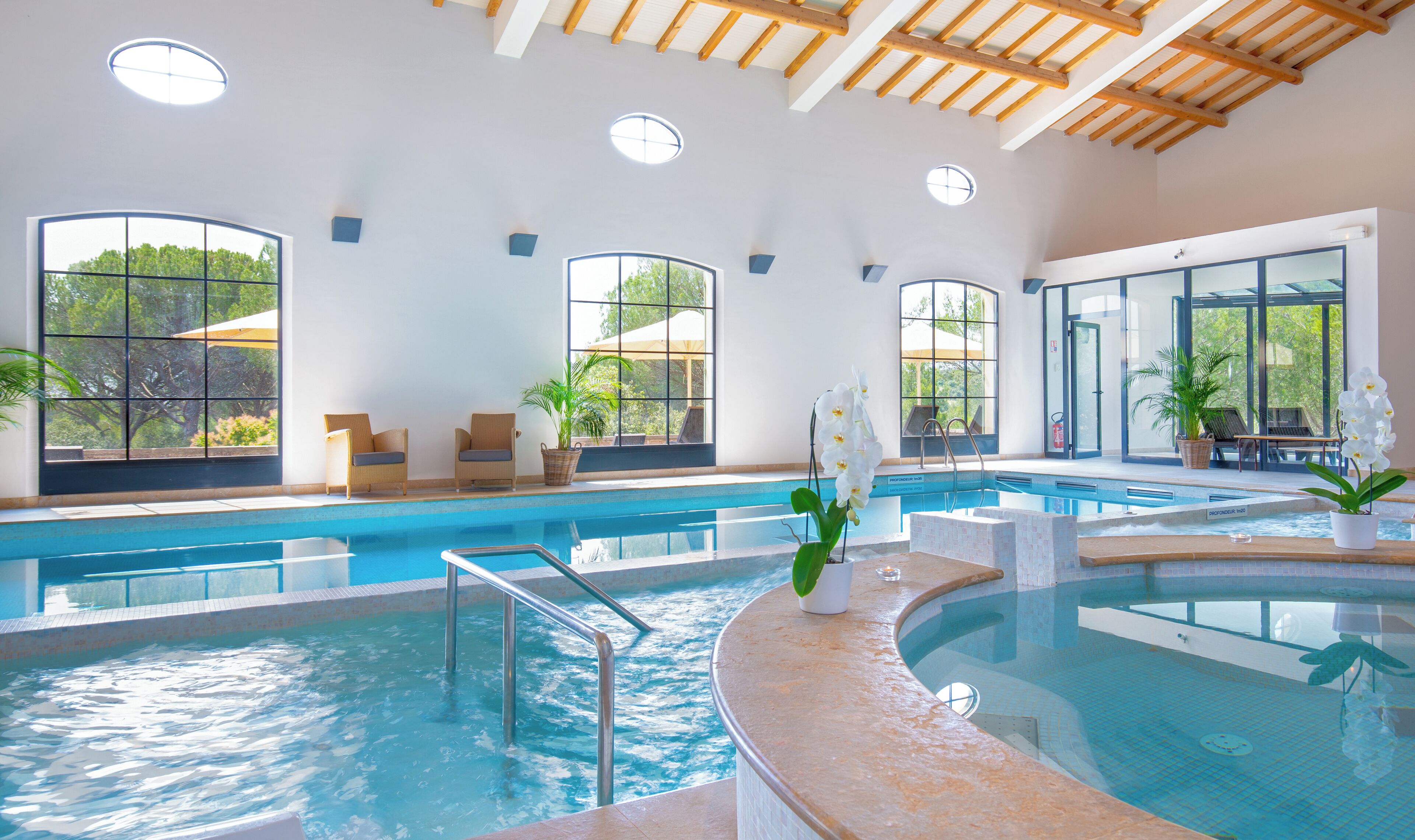 Una piscina interior con agua cristalina, vigas de madera en el techo y grandes ventanales que ofrecen vistas al verdor.
