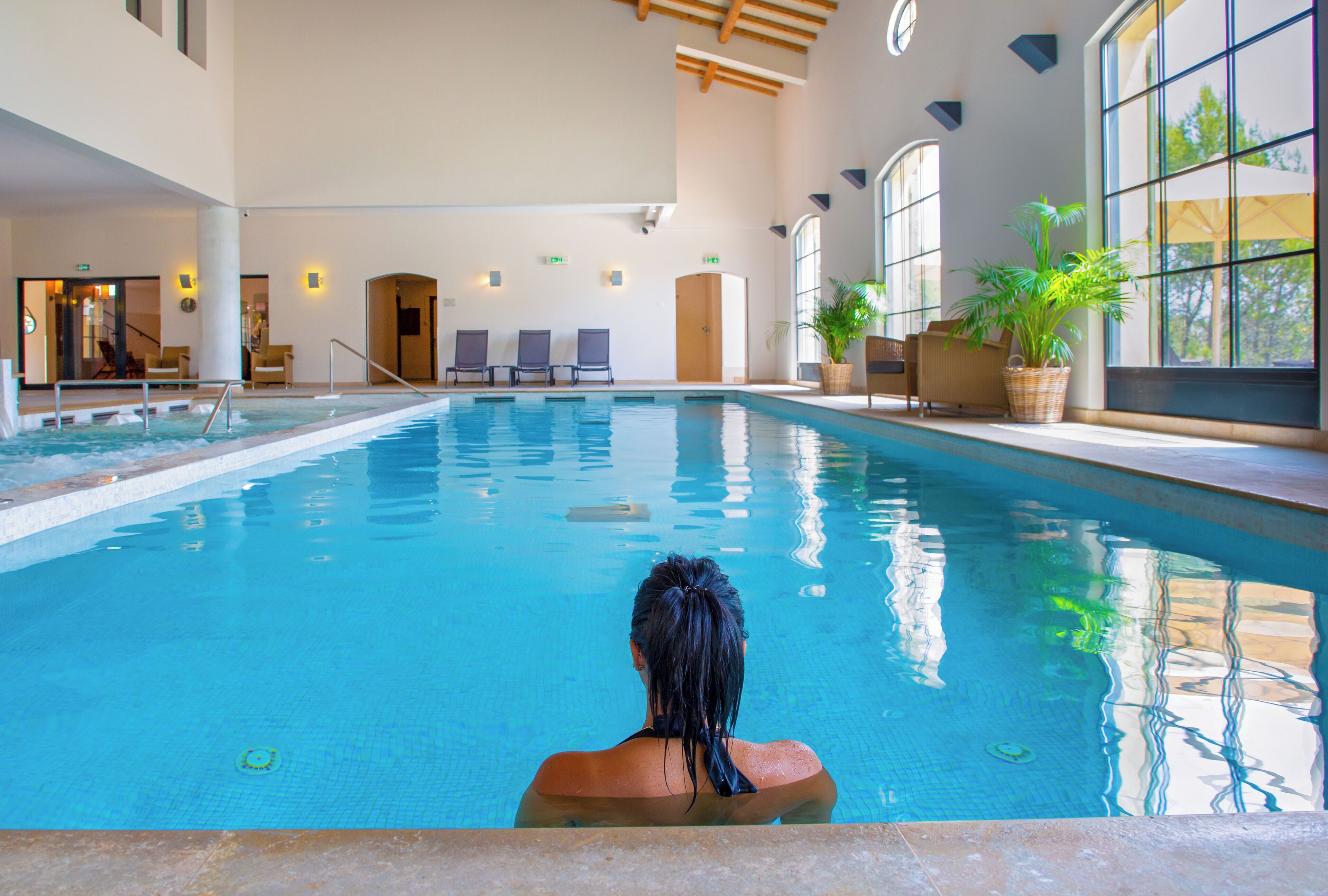 Ein großes Hallenbad mit klarem, blauem Wasser, umgeben von Liegestühlen und Topfpflanzen.