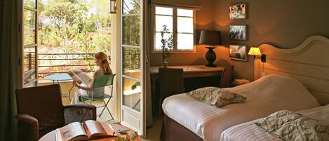 Habitación de hotel con cama, escritorio y silla. Hay una ventana con vistas a árboles y un balcón.