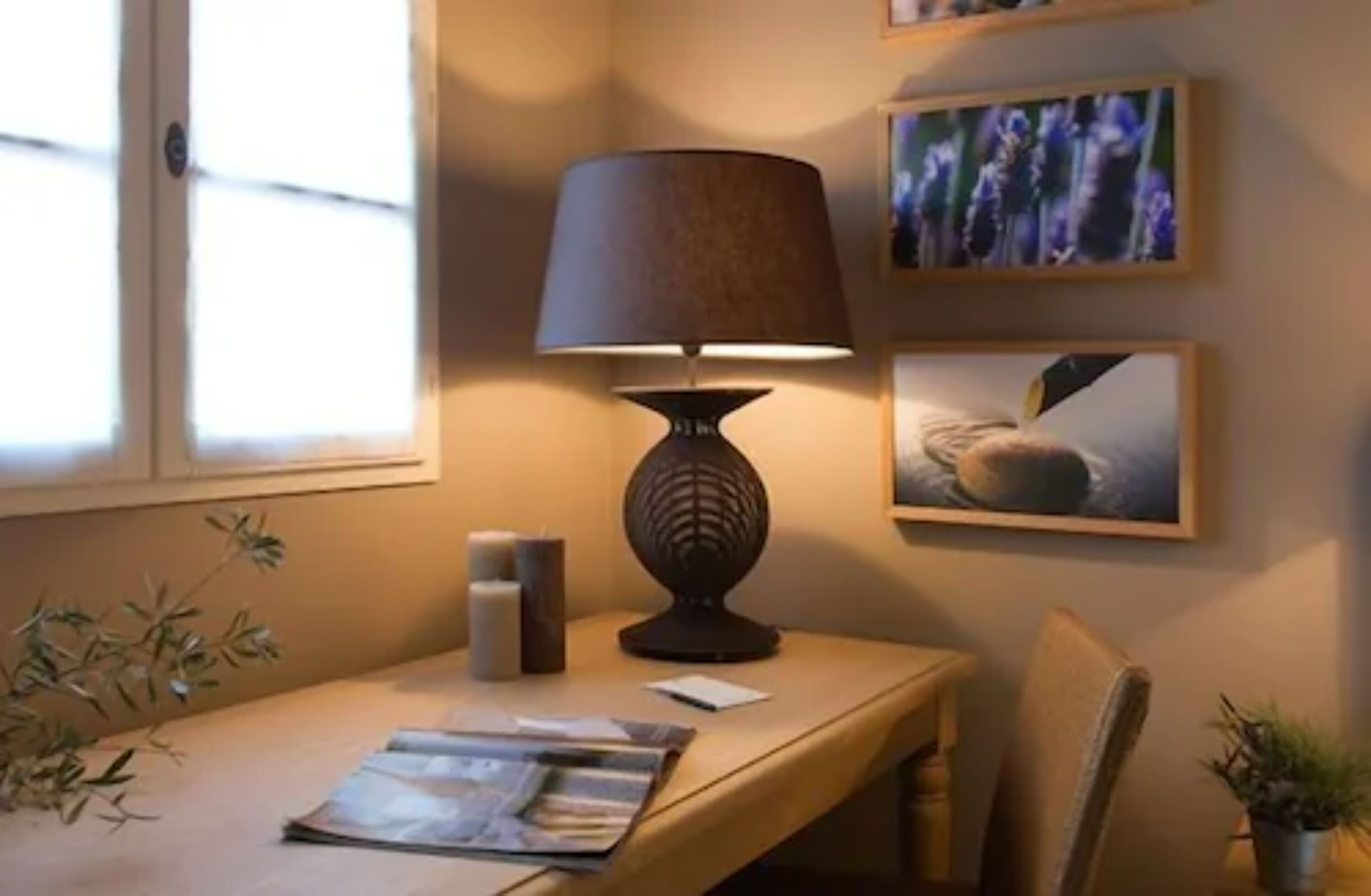 A room with a table, a lamp, a chair, a window, and wall art.
