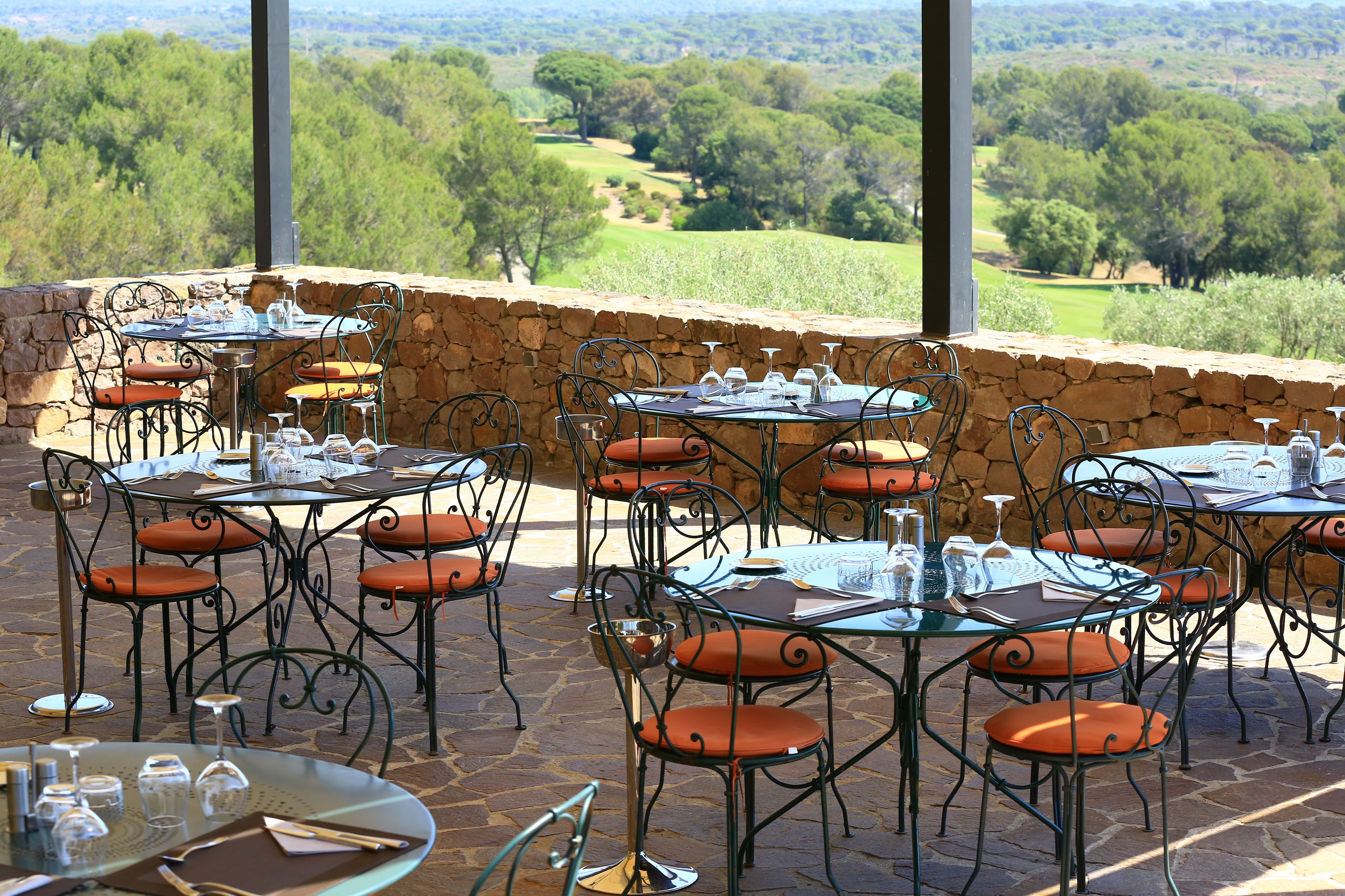 An outdoor dining area with stone walls and metal chairs, tables set with wine glasses and plates, and a view of a golf course and trees in the background.