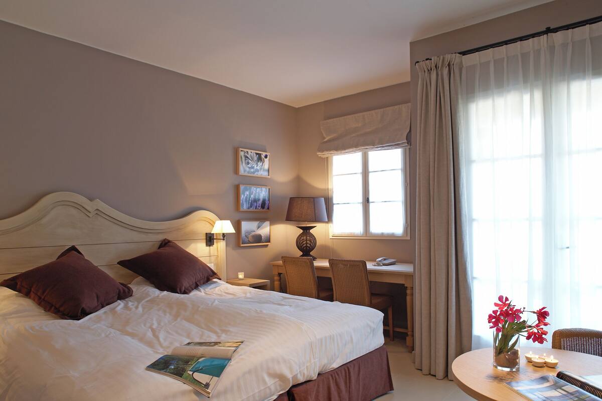 Una habitación de hotel con una cama grande, lámparas de noche, un escritorio con una silla, una ventana con cortinas y un jarrón con flores.
