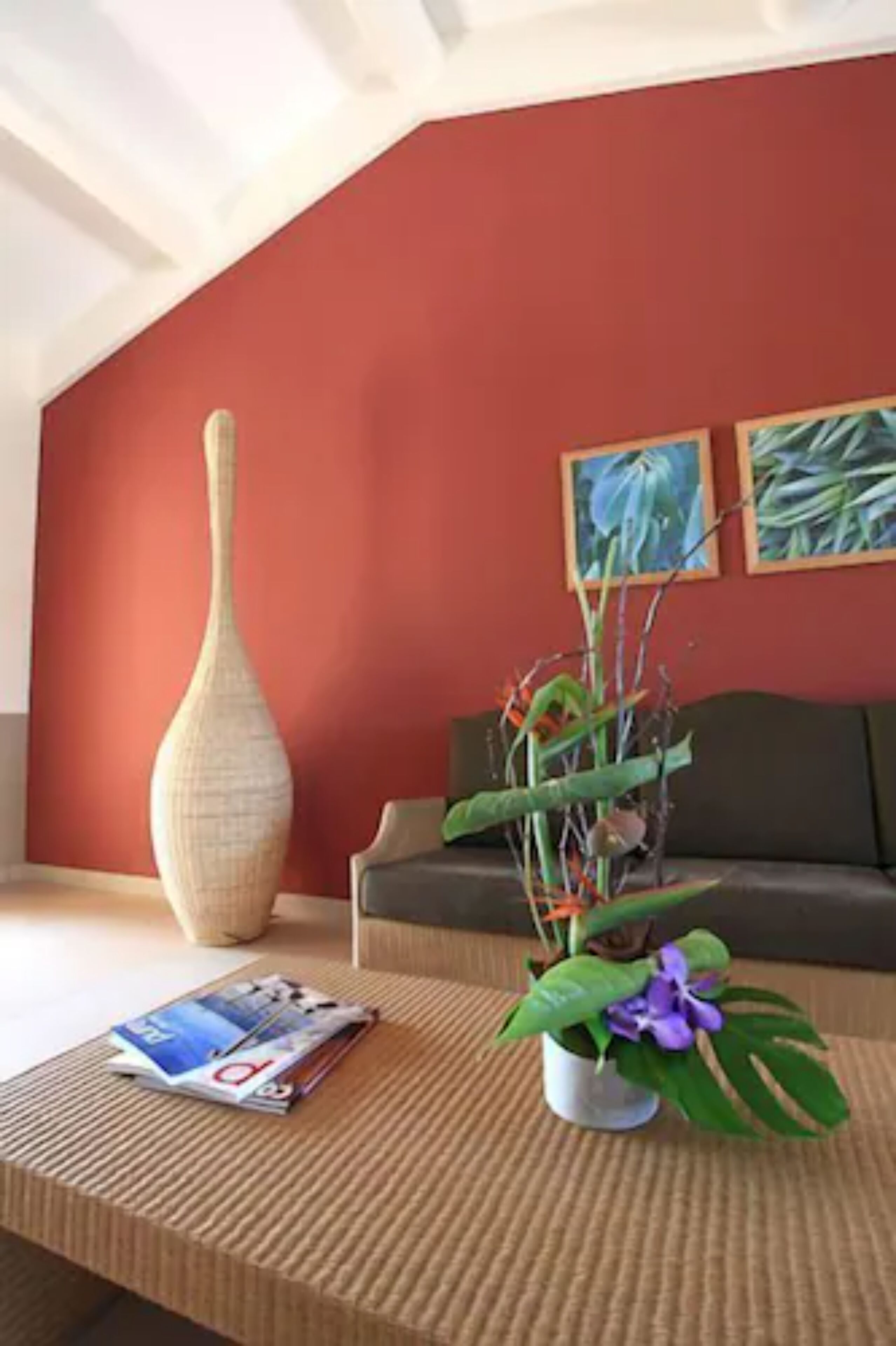 A living room with a red wall, a large vase, a potted plant, a magazine on a coffee table, and a sofa.