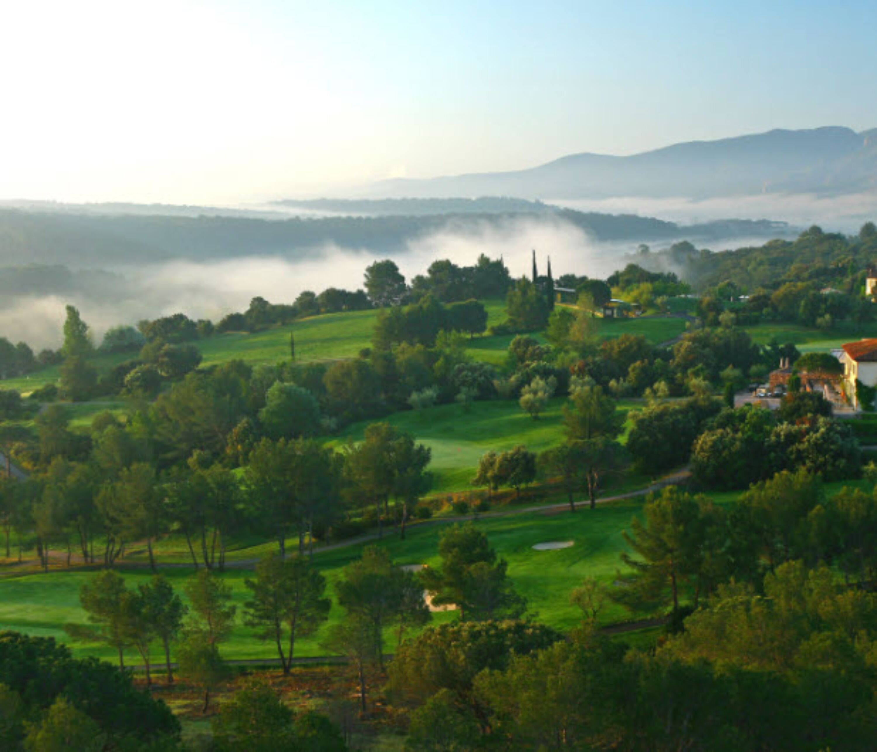 A lush green landscape with rolling hills, a golf course, and a few buildings in the distance.