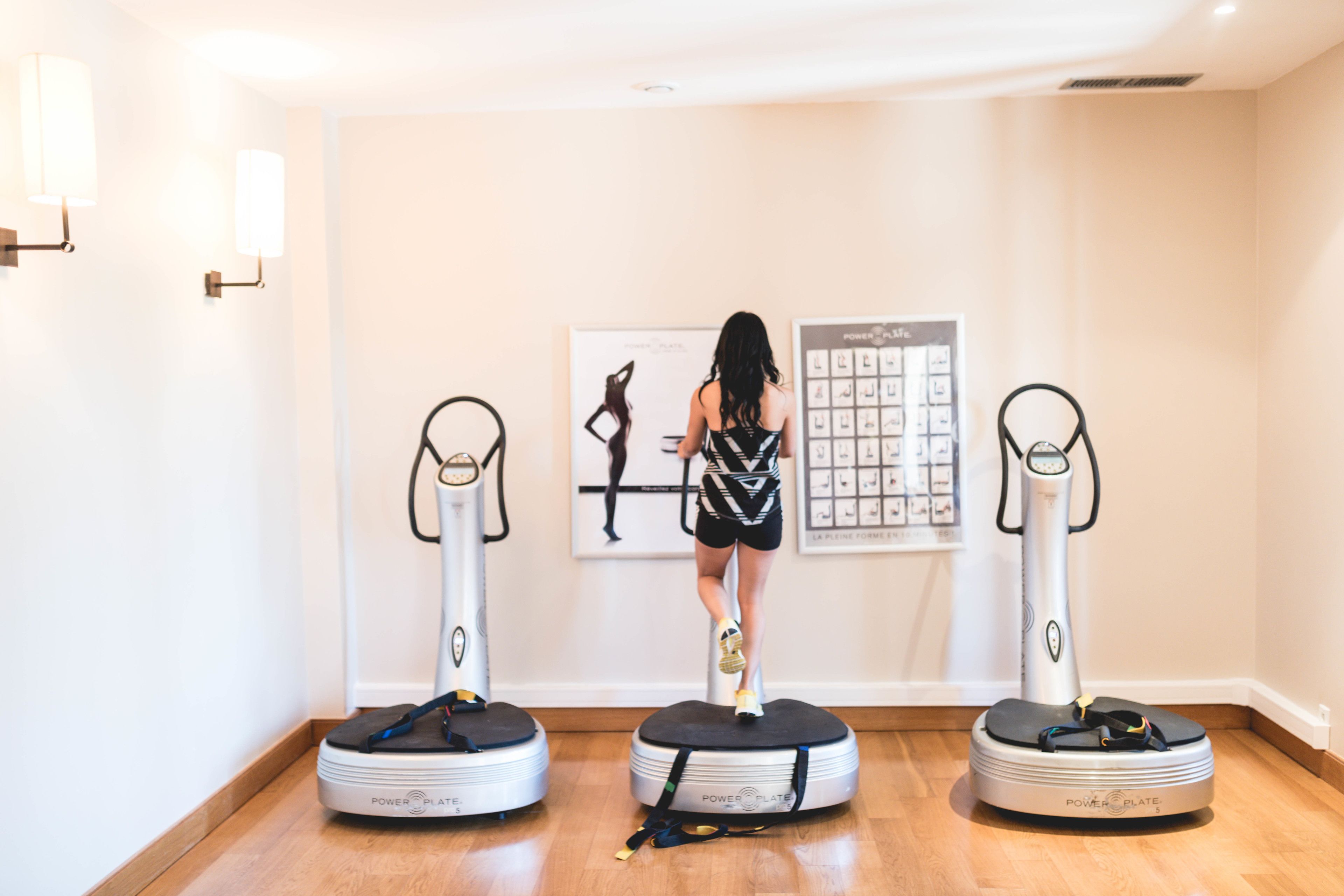 A room with three standing balance boards labeled "POWER PLATE" on the floor, a poster with a silhouette of a woman on the wall, and a light fixture on the left.