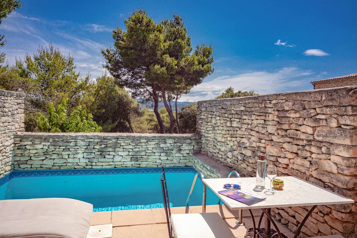 Un área de piscina con paredes de piedra, una mesa con una botella y vasos, y una silla.