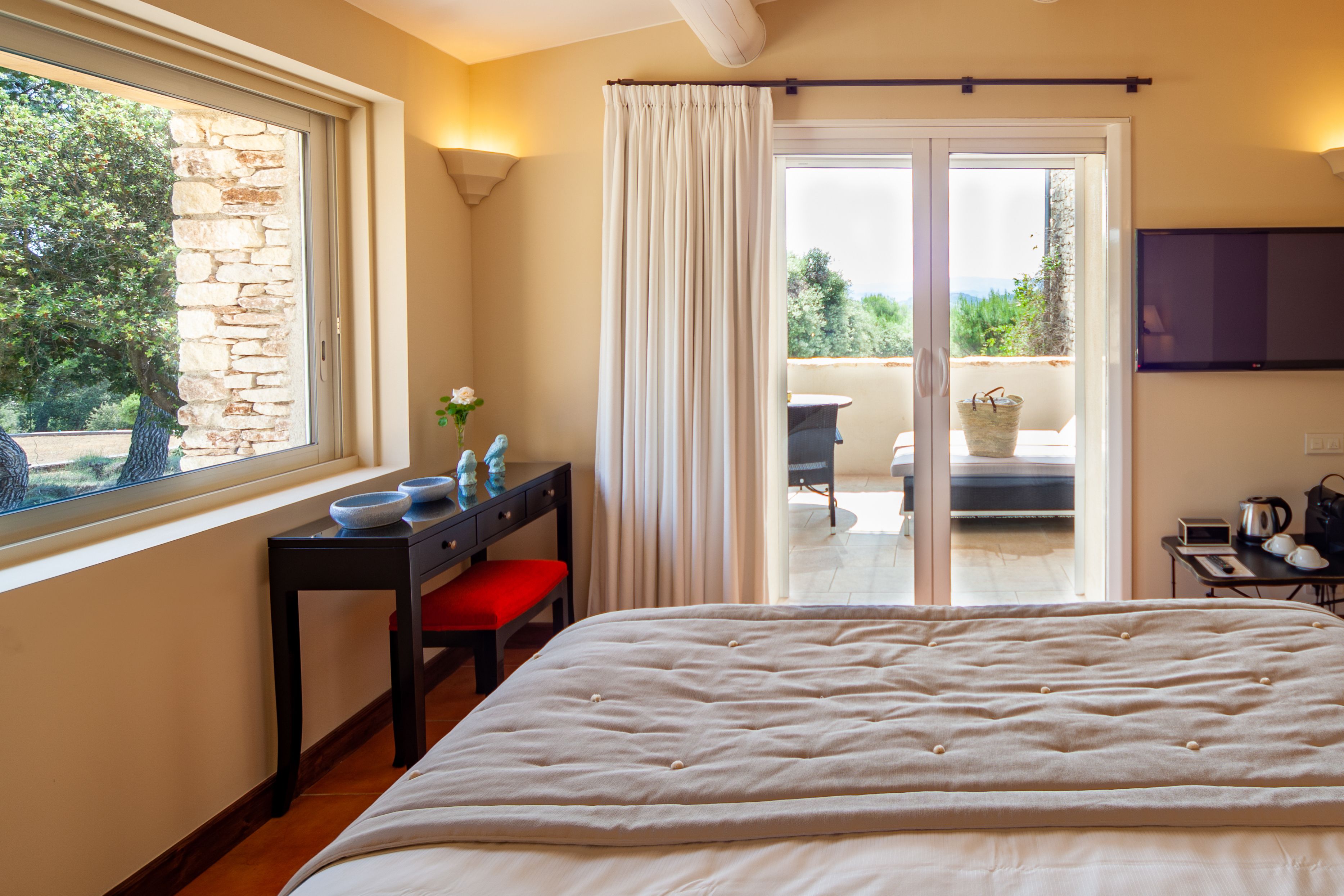 A bedroom with a large bed, a desk with a red chair, a window with a stone wall, and a balcony with a table and chairs.