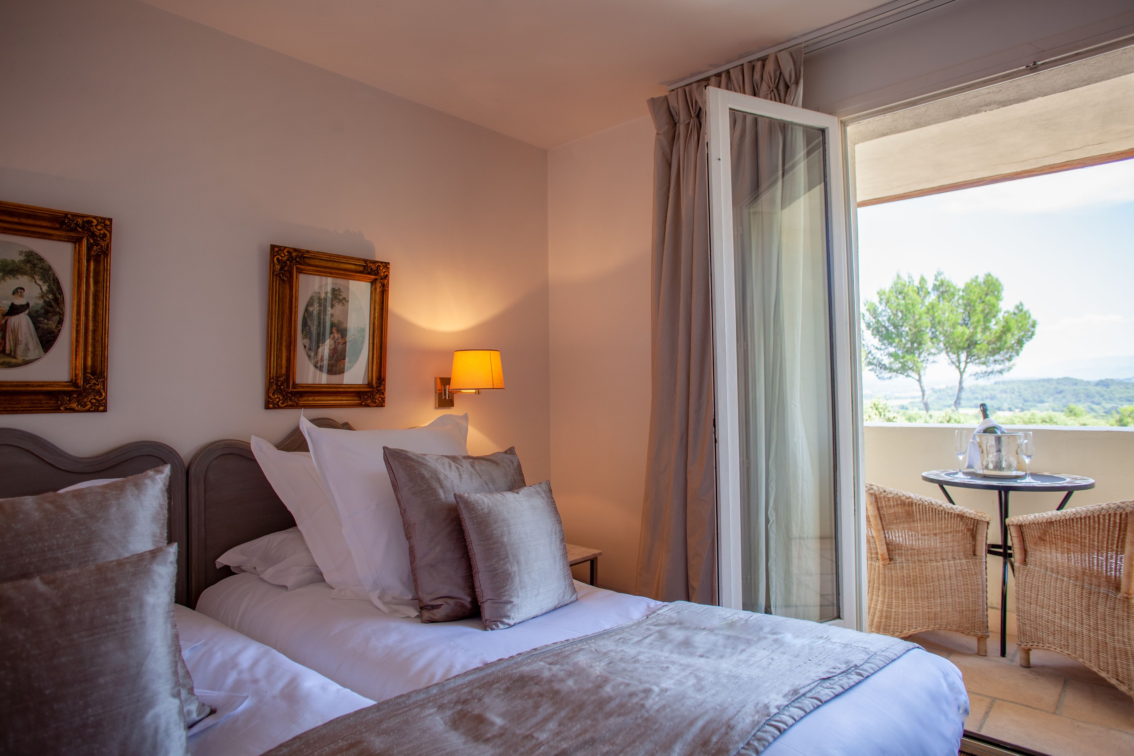 A hotel room with a bed, two pillows, a nightstand, a lamp, a window with curtains, and a balcony with wicker chairs and a table.