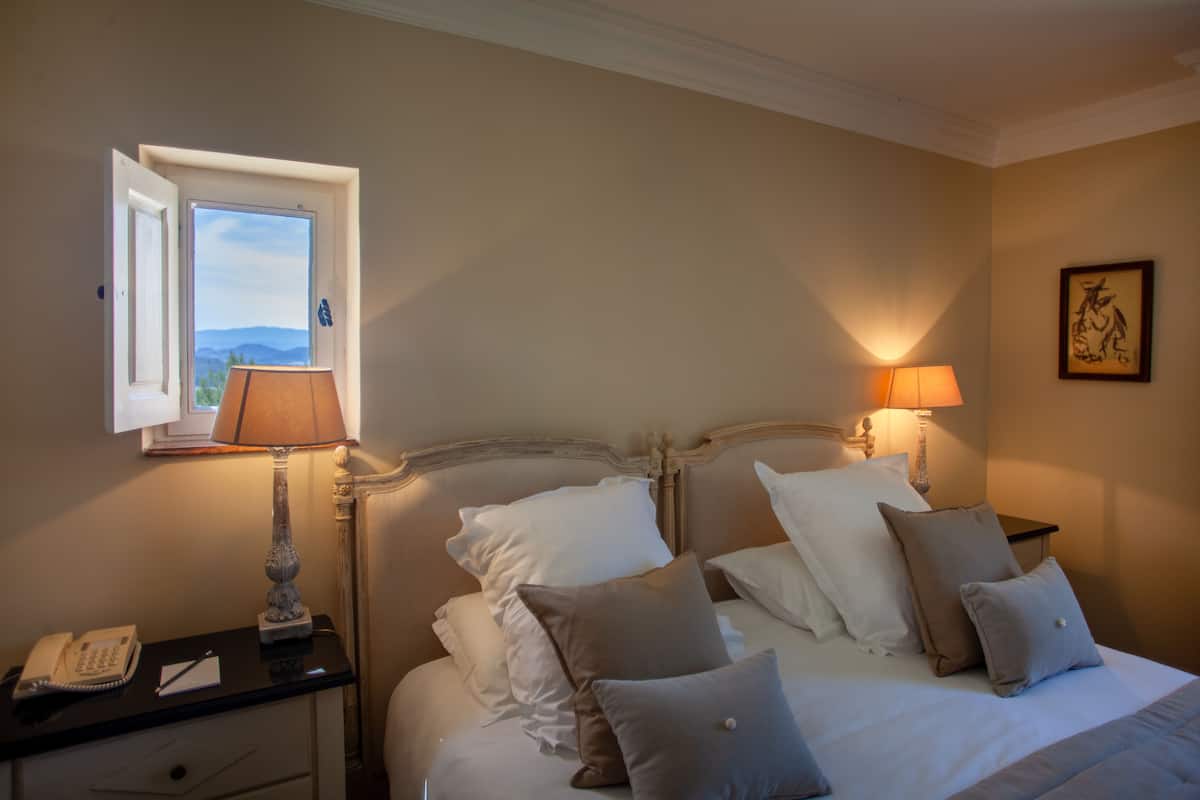Habitación de hotel con cama, lámparas de noche, mesita de noche con teléfono y ventana con vistas a las montañas.