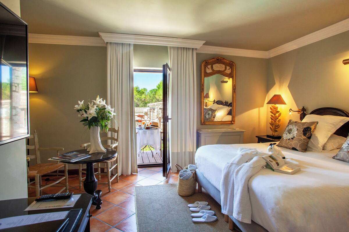 Habitación de hotel con cama, escritorio con lámpara, una silla, mesa con un jarrón de flores y vista al exterior.