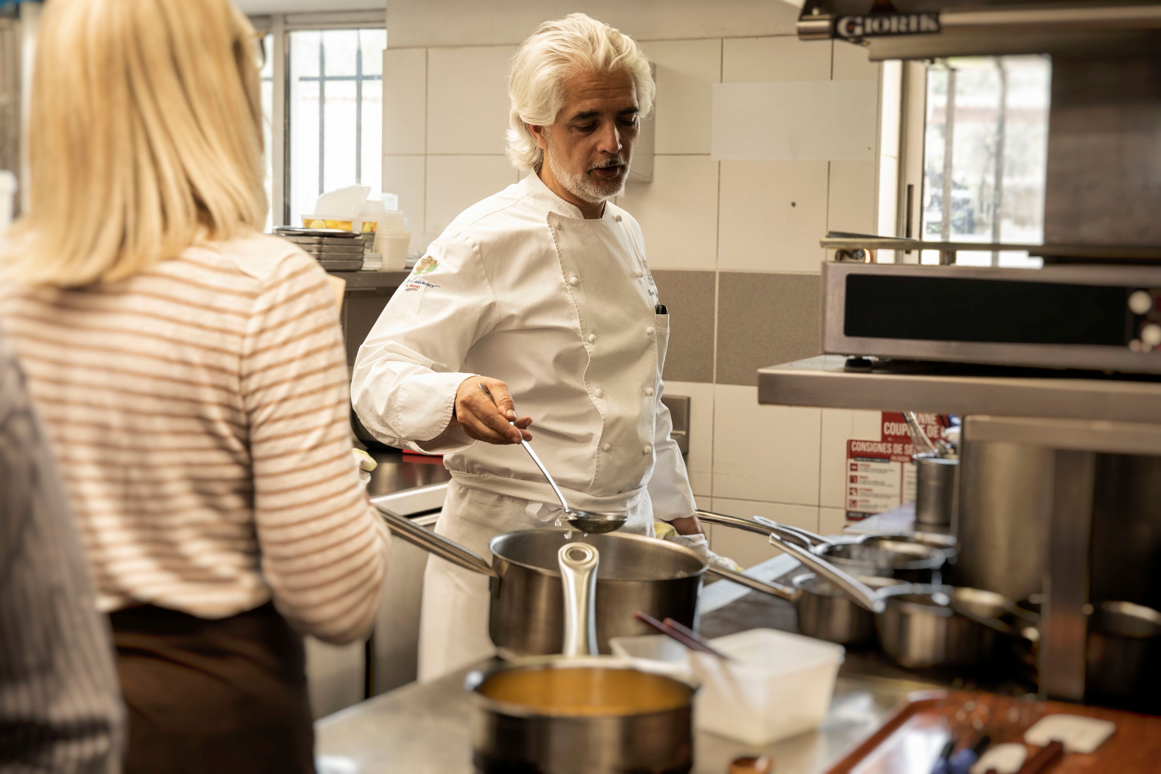 A chef in a kitchen with stainless steel appliances and a window in the background.