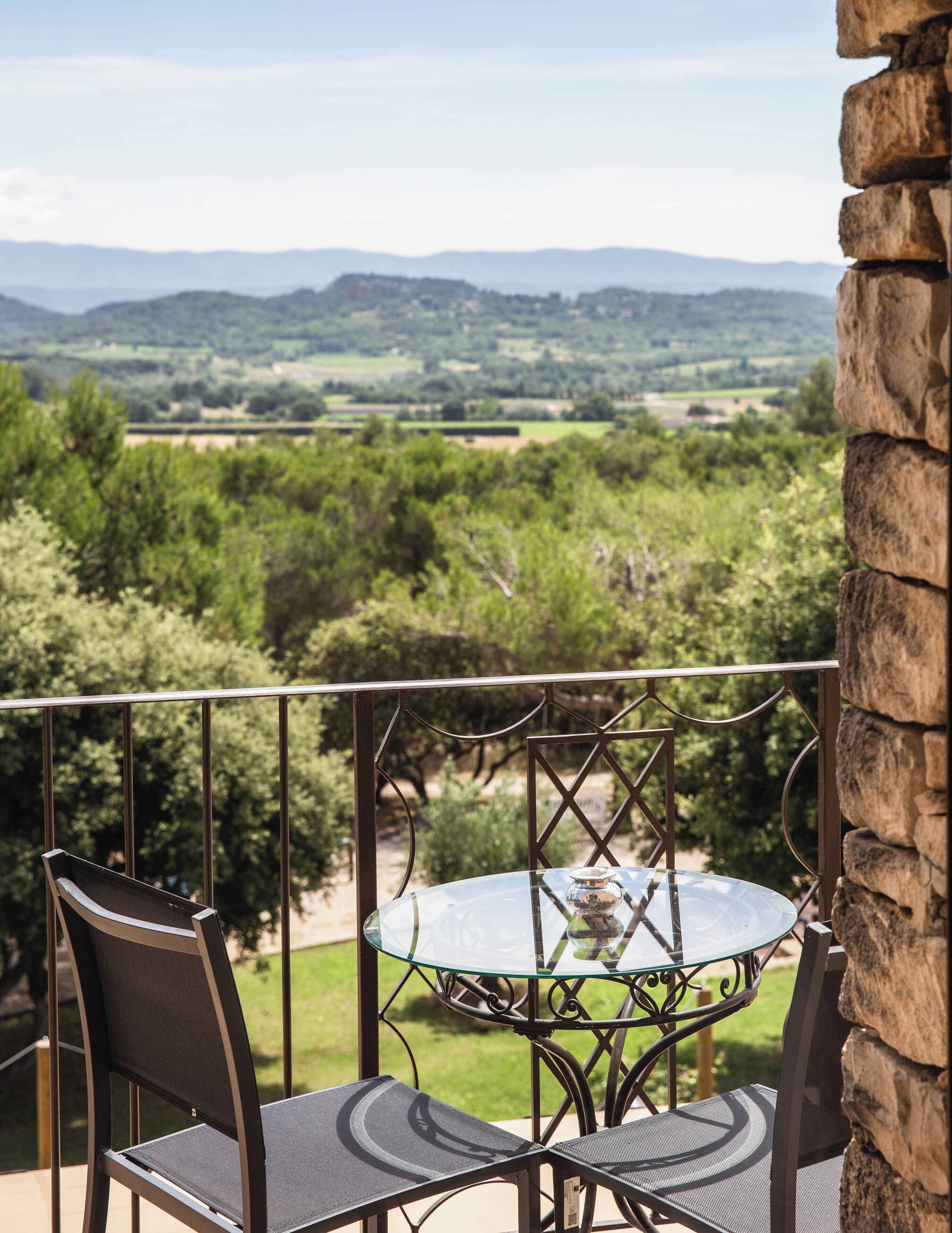 Een balkon met een glazen tafel en stoelen, met uitzicht op een schilderachtig landschap met bomen en heuvels.