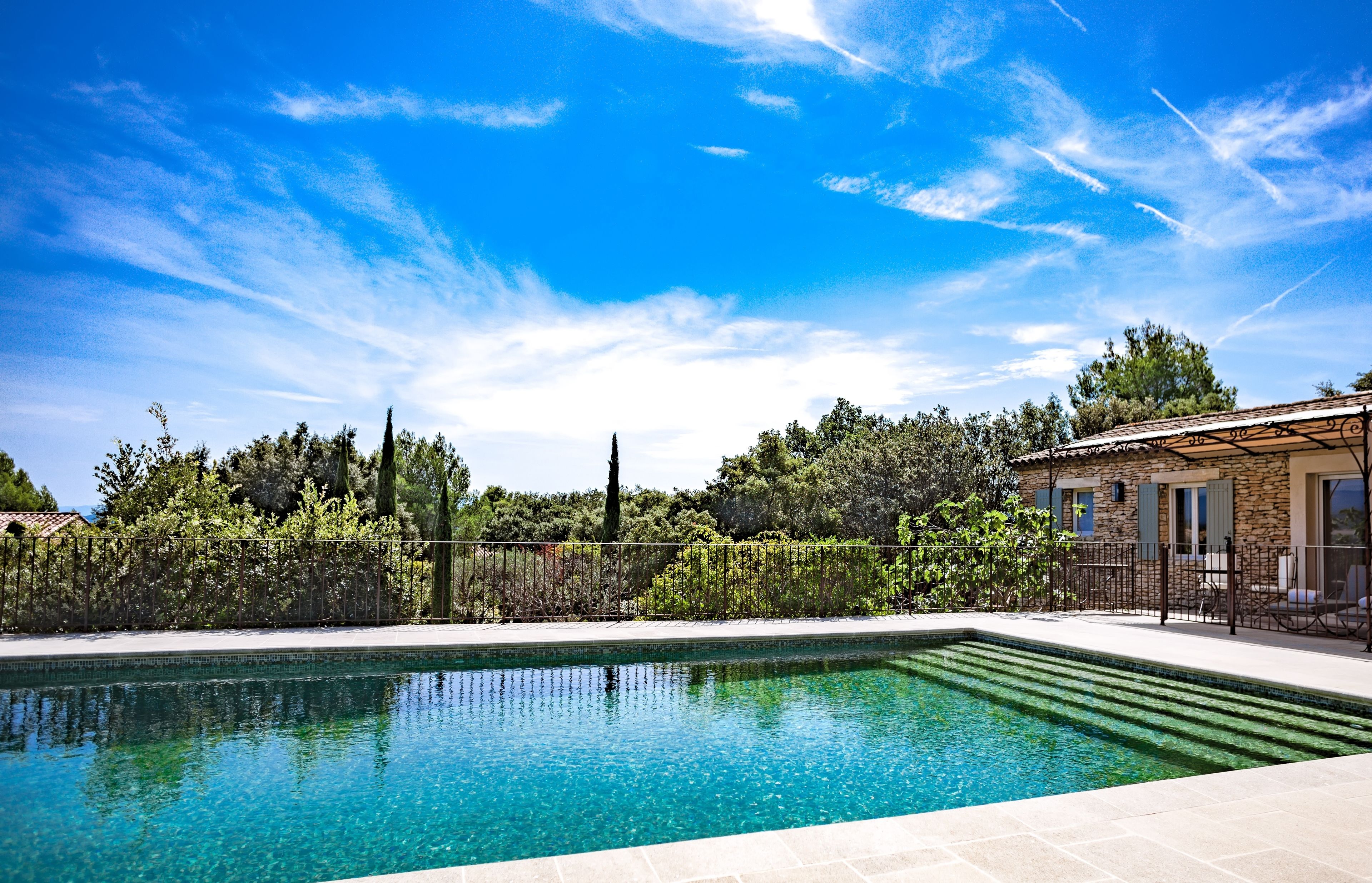 A rectangular swimming pool with clear blue water, surrounded by a stone patio and a fence.