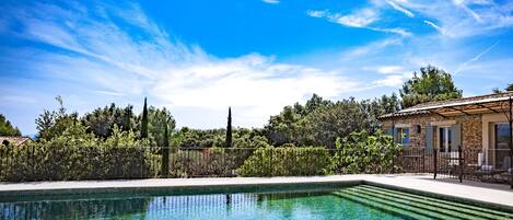 A rectangular swimming pool with clear blue water, surrounded by a stone patio and a fence.