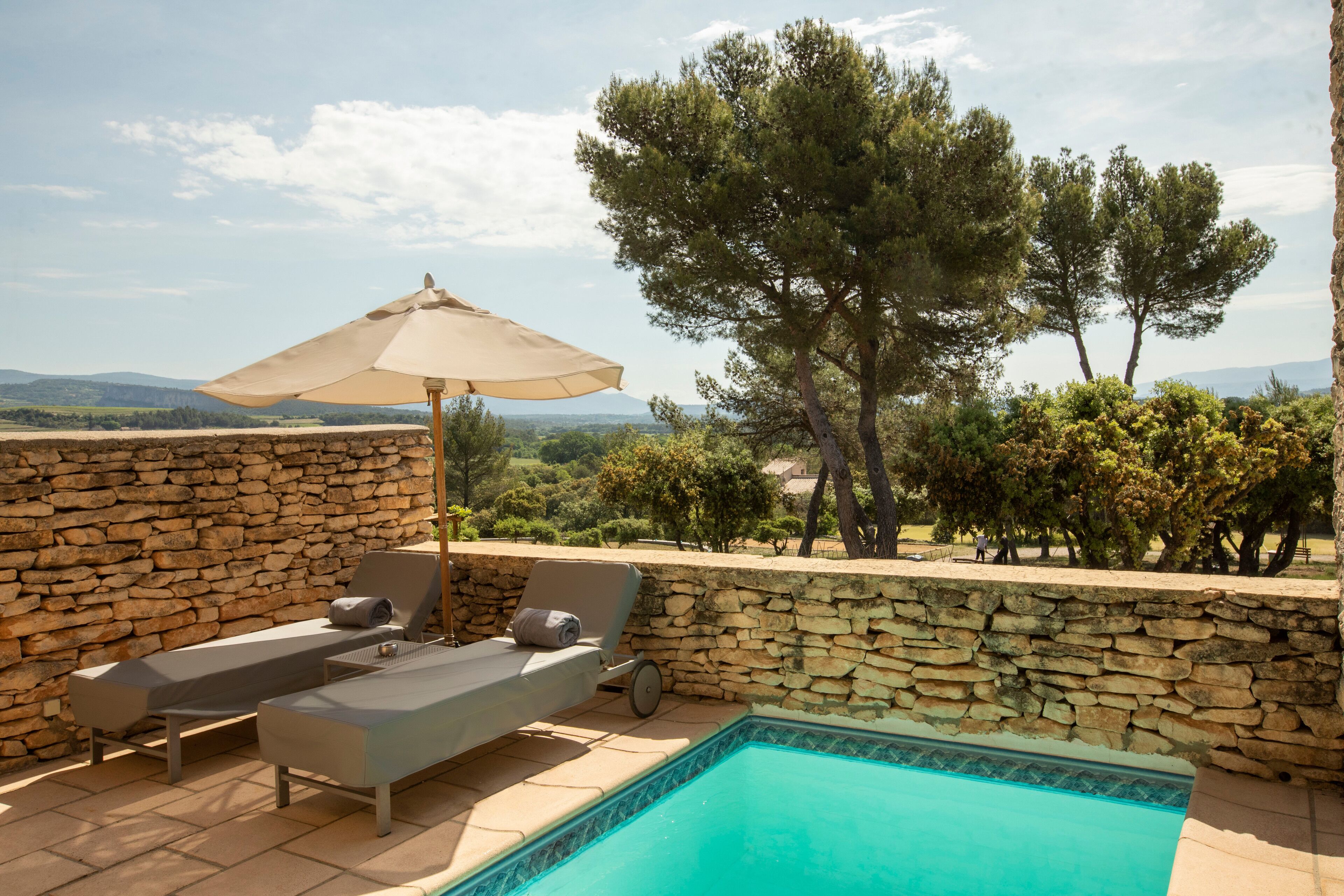 Un espace aménagé au bord de la piscine, avec des chaises longues et un auvent, entouré de murs de pierre et d’arbres, offrant une vue sur un paysage pittoresque.
