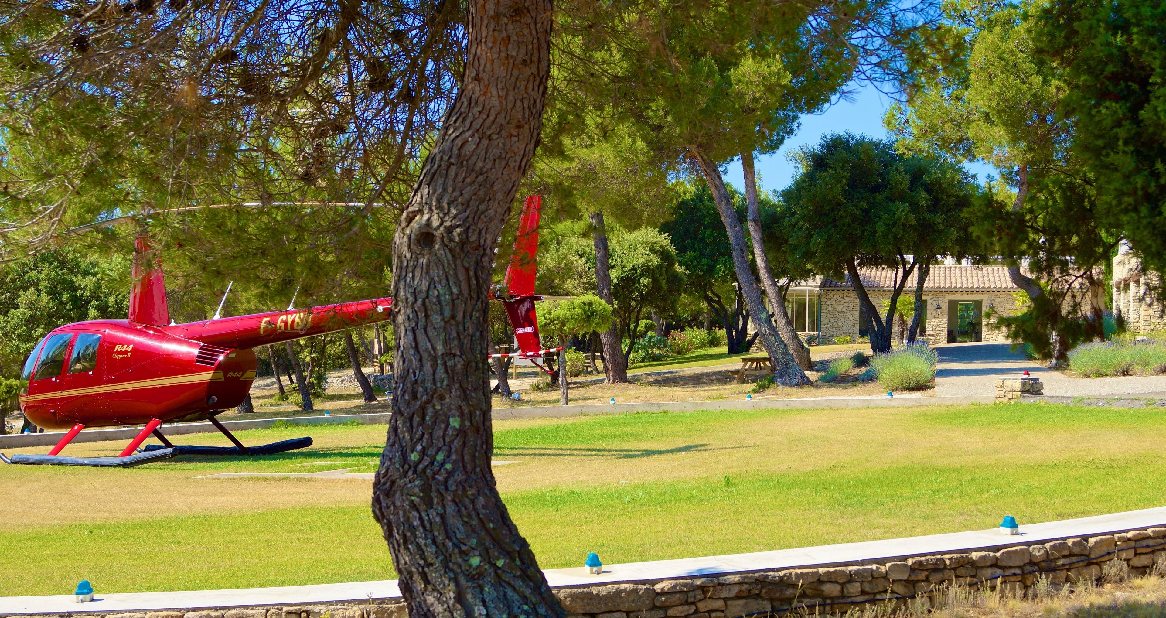 A red helicopter parked in a grassy area with trees and a stone building in the background.