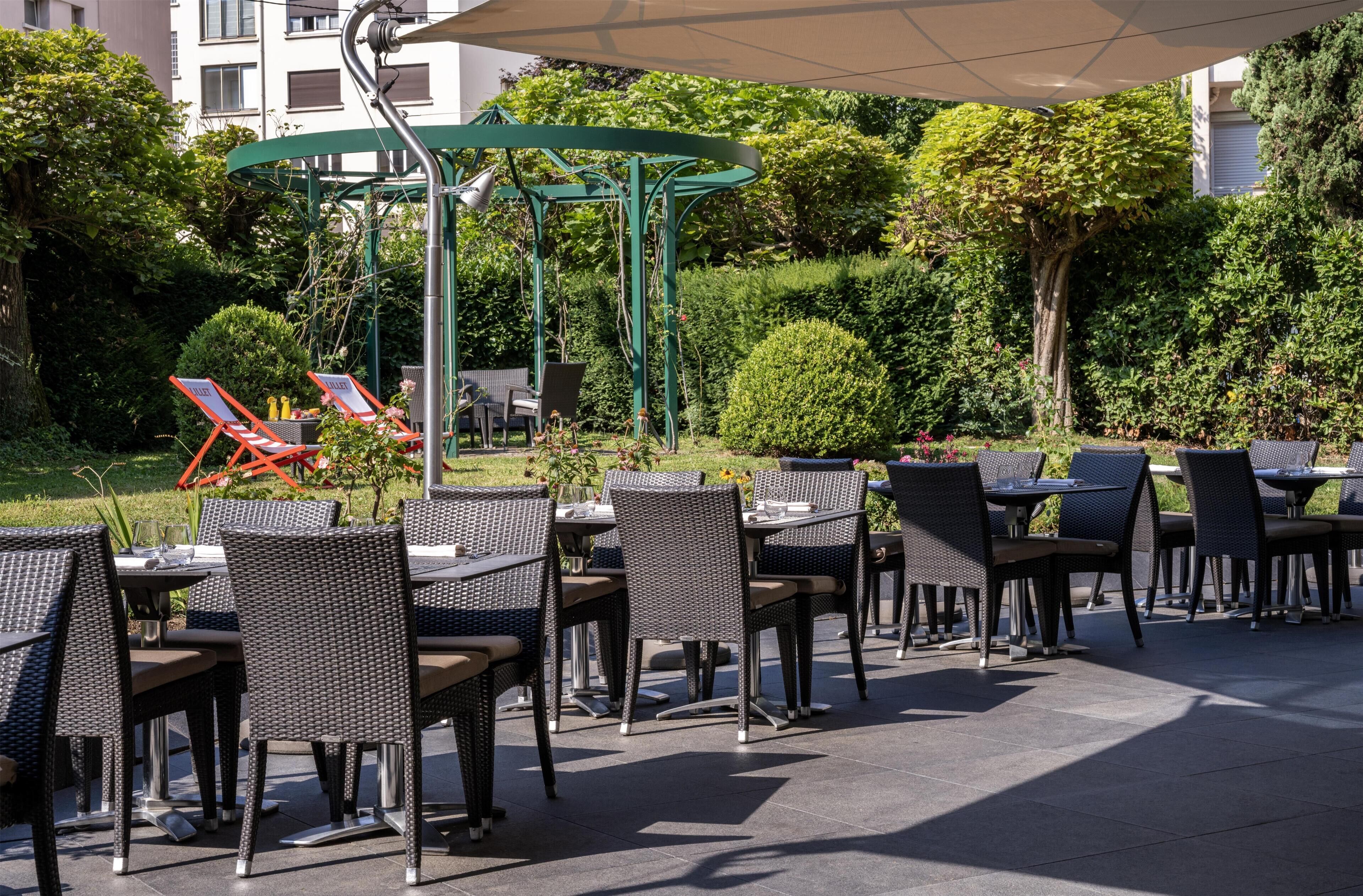 An outdoor seating area with wicker chairs and tables, a pergola structure, and a backdrop of greenery and buildings.