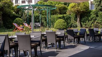 An outdoor seating area with wicker chairs and tables, a pergola structure, and a backdrop of greenery and buildings.