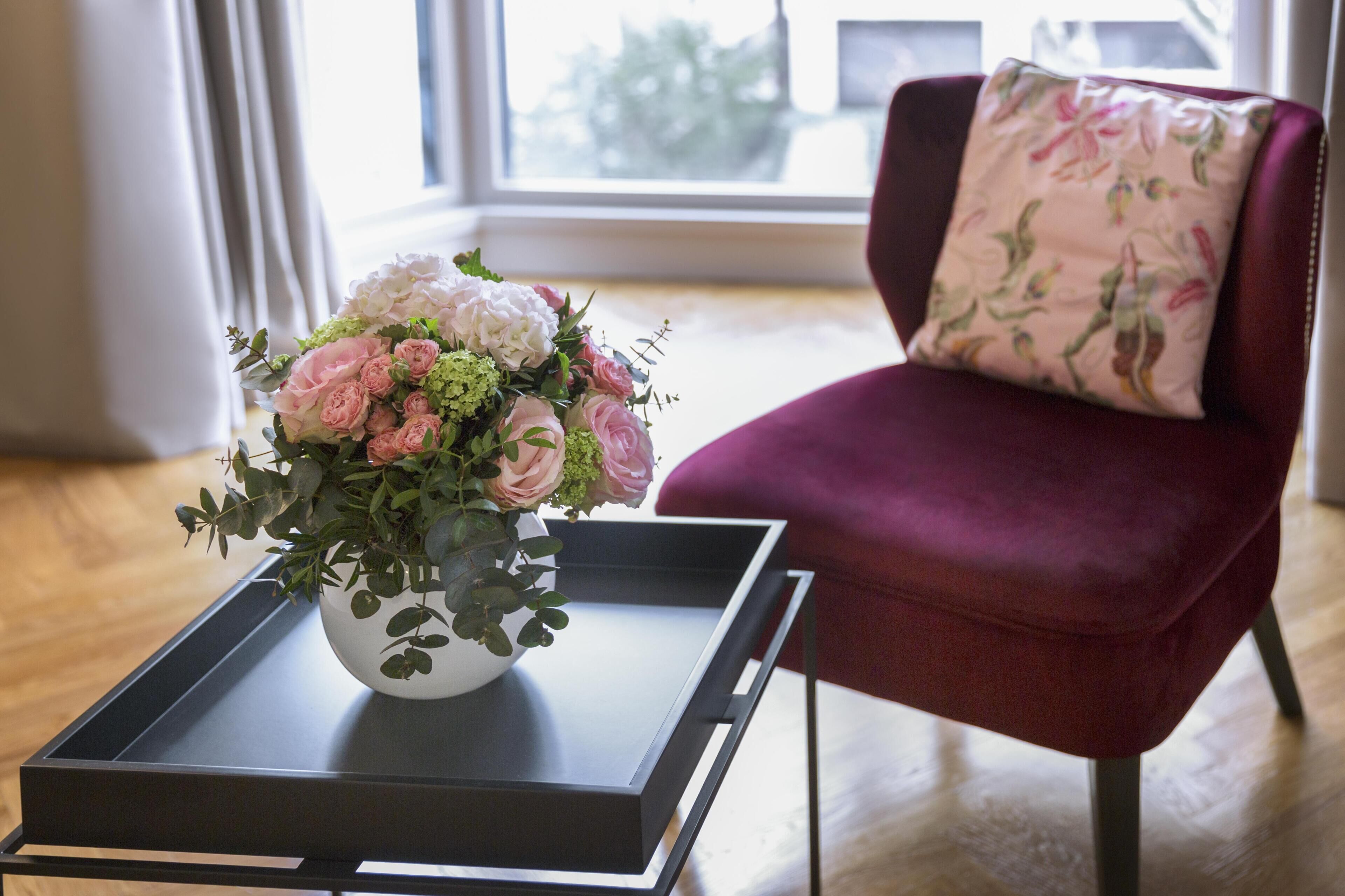 A room with a floral arrangement on a tray table, a red chair with a floral pillow, and a window with a view of trees.