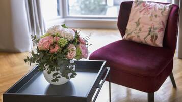 A room with a floral arrangement on a tray table, a red chair with a floral pillow, and a window with a view of trees.