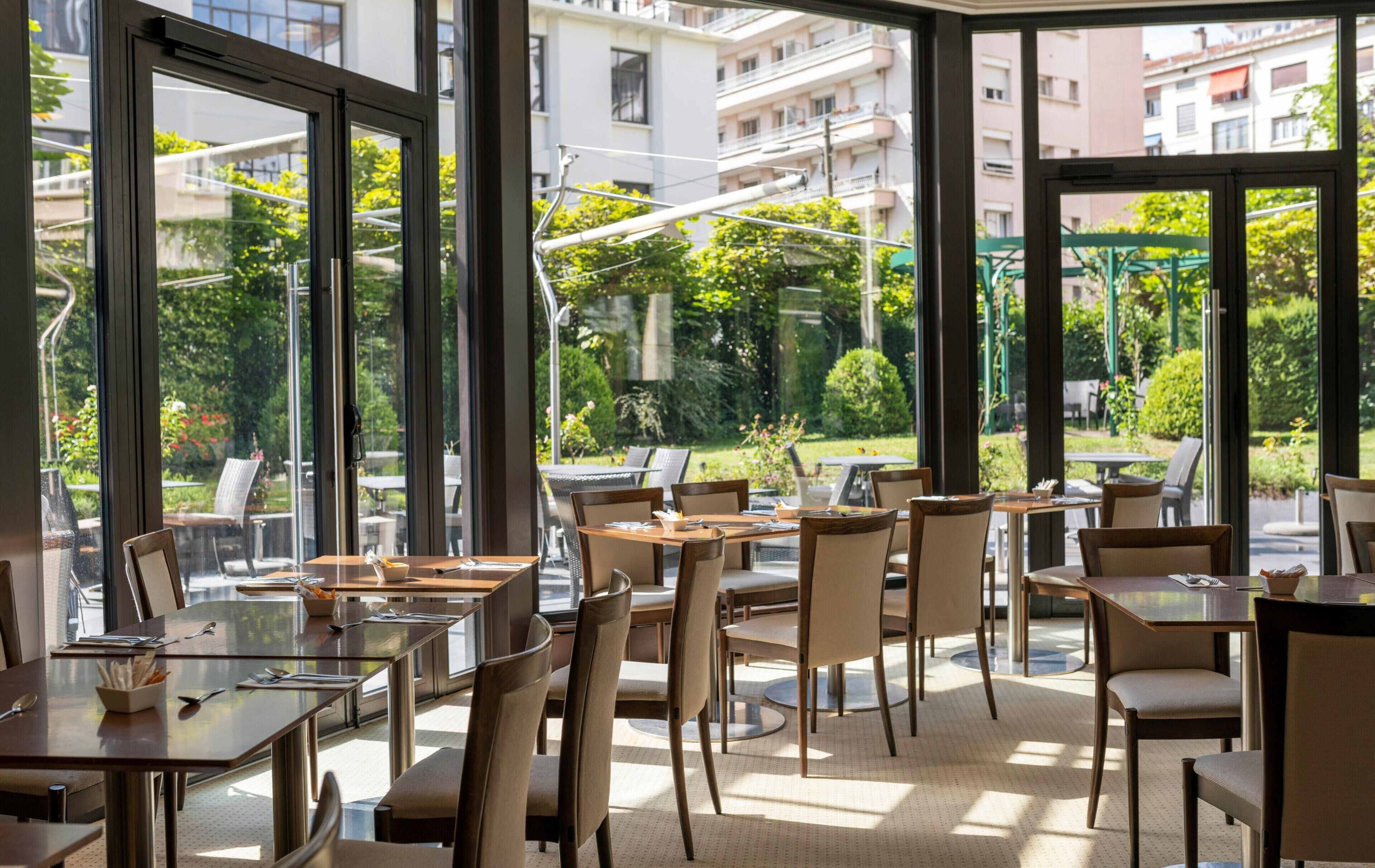 An indoor dining area with tables and chairs, large windows, and a view of greenery and buildings outside.