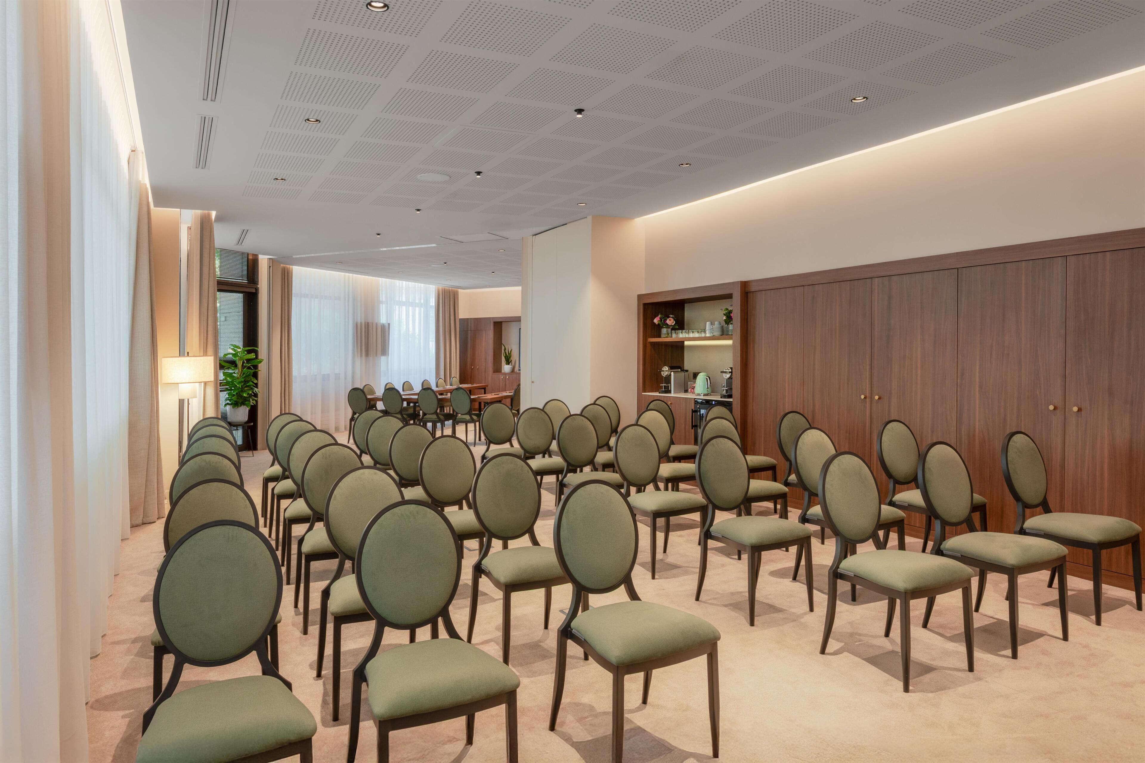 A conference room with rows of chairs, a wooden cabinet with glassware, and a ceiling with a grid pattern.