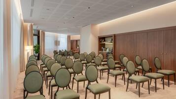 A conference room with rows of chairs, a wooden cabinet with glassware, and a ceiling with a grid pattern.