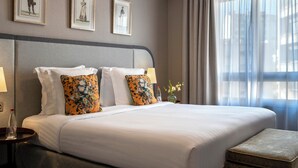 Habitación de hotel con una cama grande, dos mesitas de noche, un velador, una ventana con cortinas traslúcidas y tres cuadros en la pared.