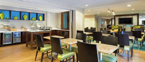 A modern restaurant with multiple dining tables and chairs, a kitchen area, and a television screen.