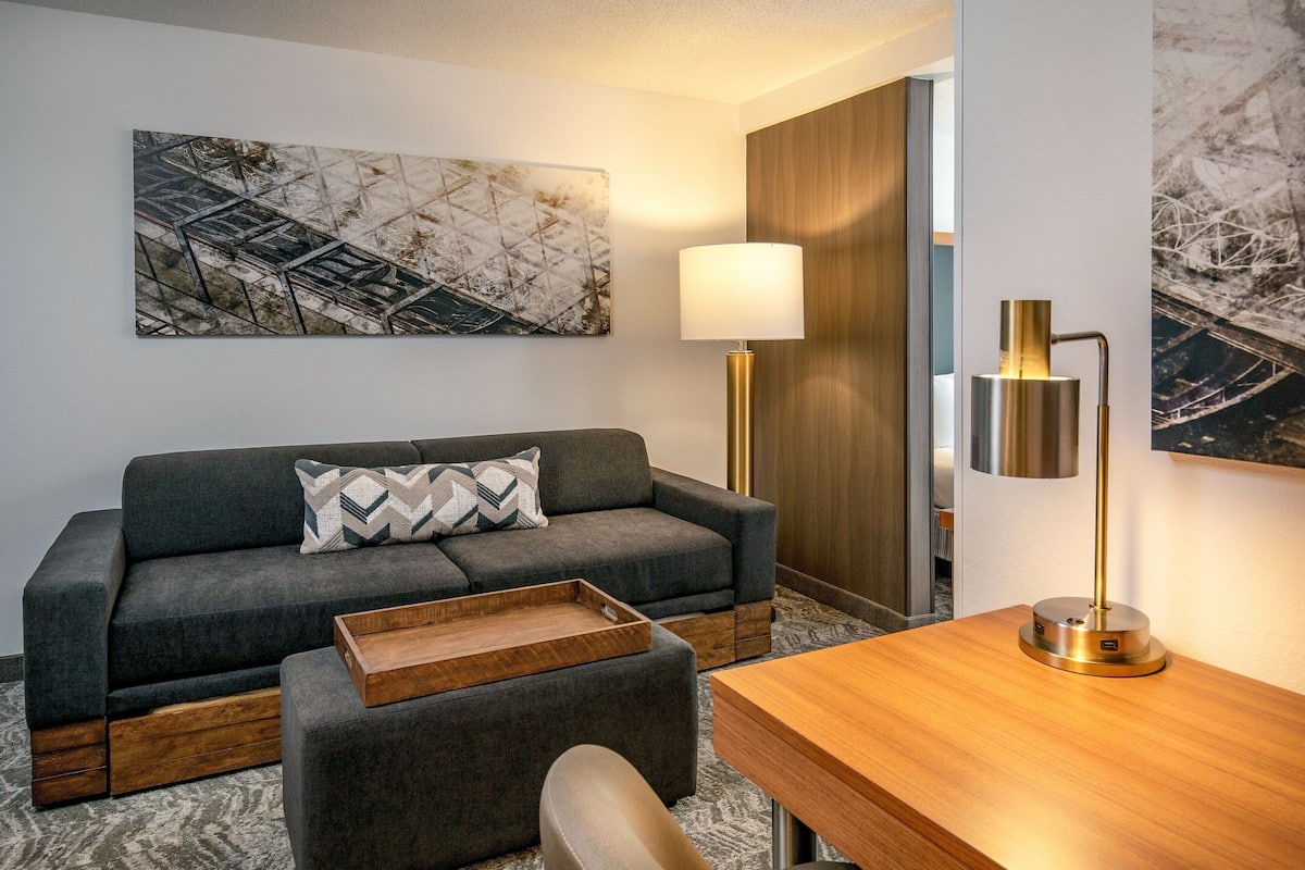 A modern hotel room with a grey sofa, a wooden side table, a desk with a lamp, and abstract artwork on the walls.