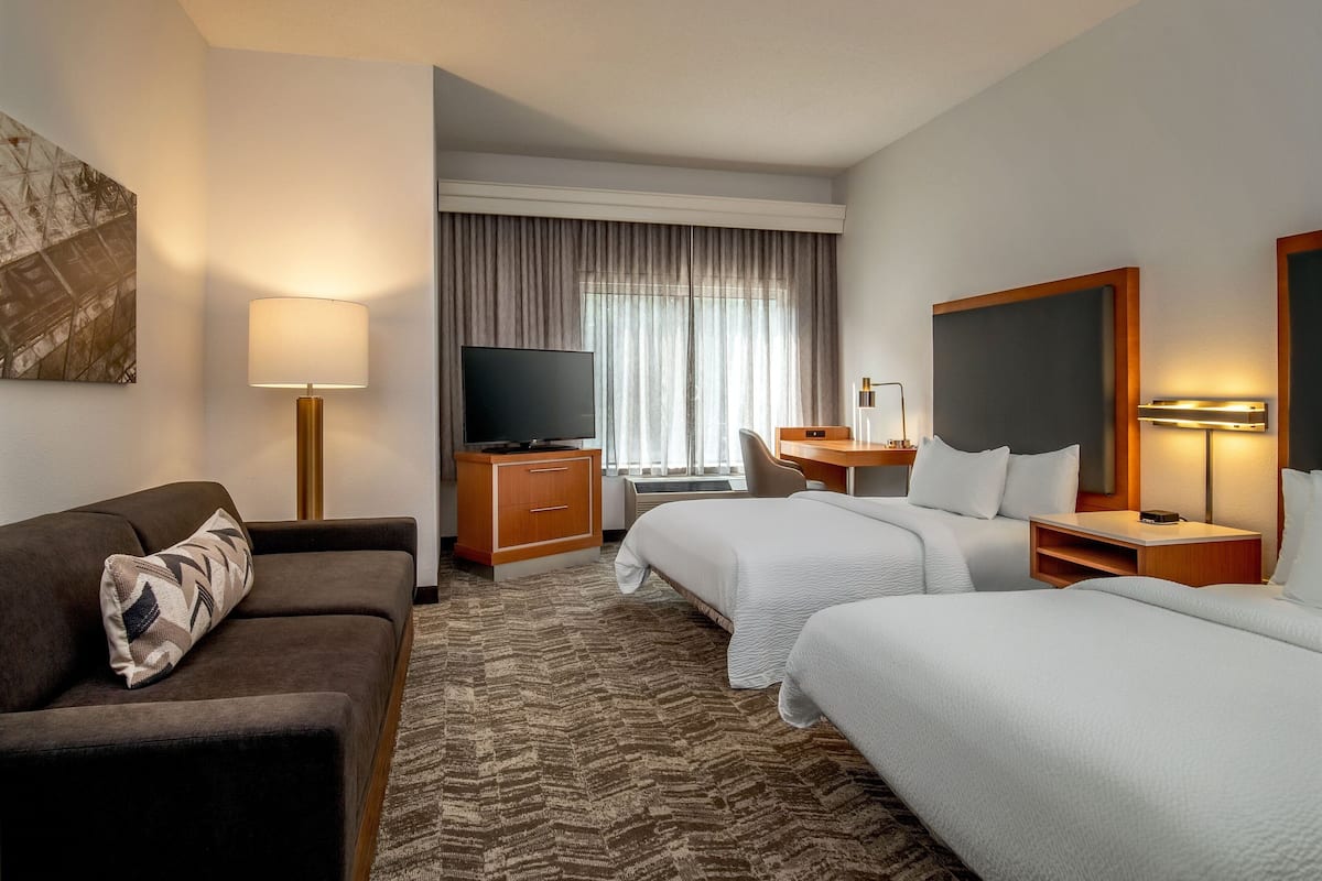 A hotel room with a sofa, two beds, a TV, a desk, and a window with curtains.