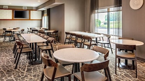 Un salón de conferencias de hotel con varias mesas y sillas, un televisor de pantalla plana grande montado en la pared, y un amplio ventanal con vistas al exterior.