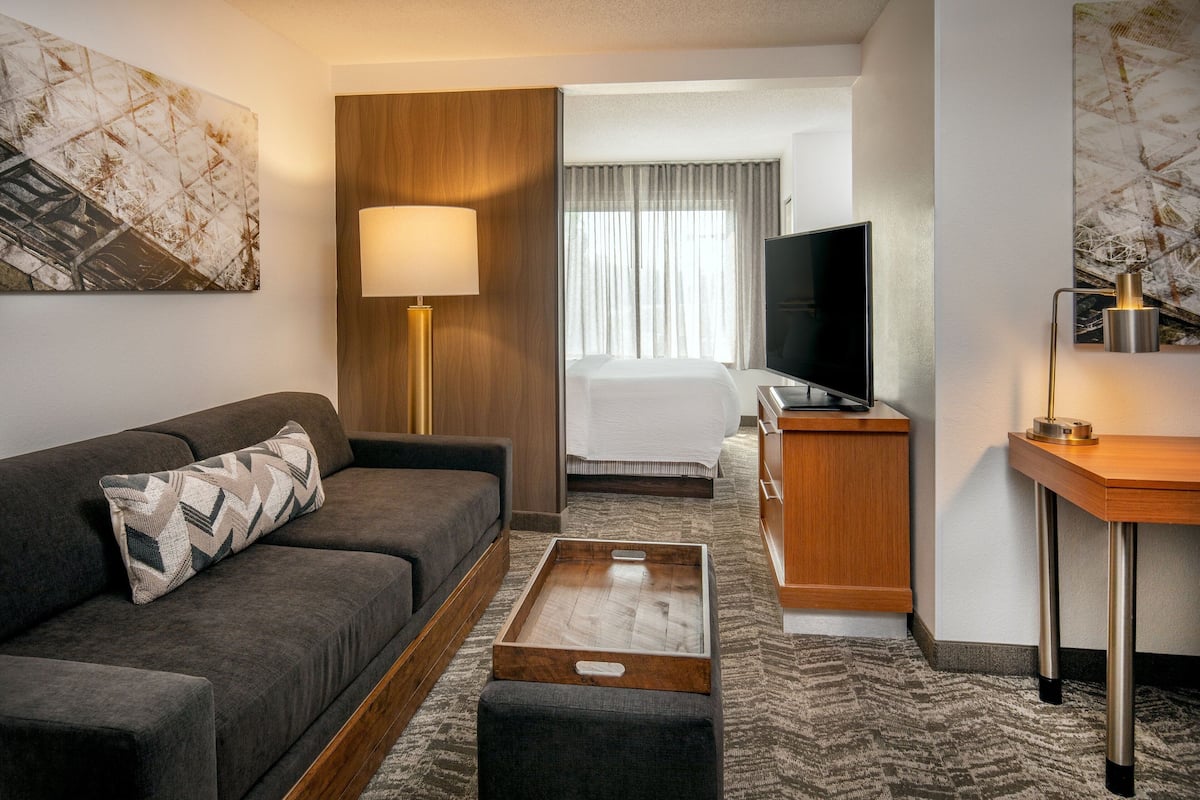 A modern hotel room with a sofa, a wooden coffee table, a desk, a television, and a bed.