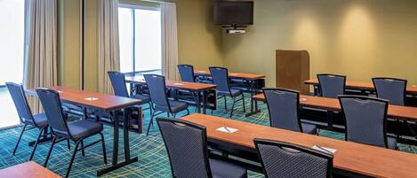 Un salón de conferencias con varias mesas y sillas, un amplio ventanal con cortinas y un televisor de pantalla plana montado en la pared.
