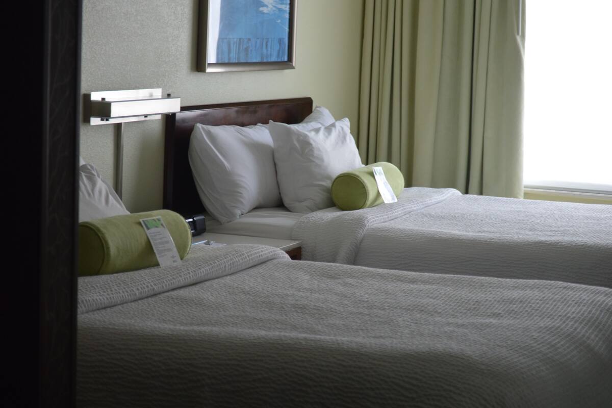 Ein Hotelzimmer mit zwei Betten, jeweils mit weißer Bettwäsche und grünen Kissen. Über den Betten hängt ein gerahmtes Bild und ein Fenster mit Vorhängen.