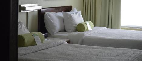 Habitación de hotel con dos camas, cada una con ropa blanca y almohadas verdes. Hay un cuadro enmarcado en la pared sobre las camas y una ventana con cortinas.