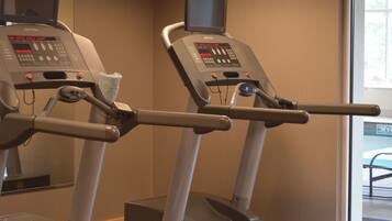 Two exercise machines with digital displays and control panels, positioned in a room with a window and a door in the background.