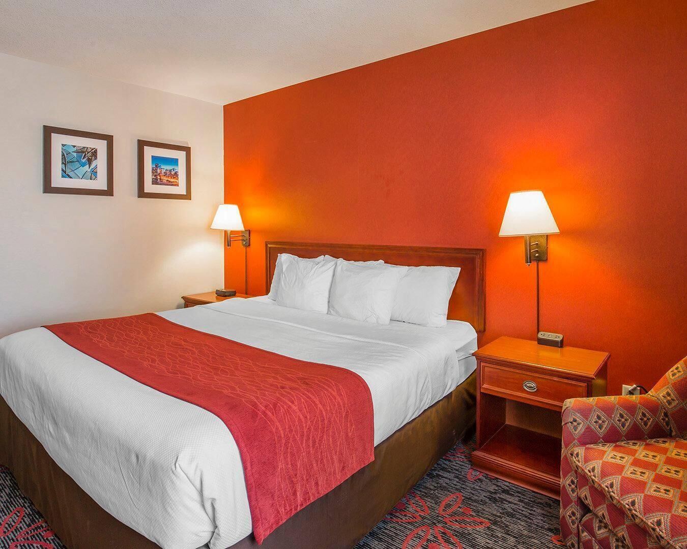 Une chambre d’hôtel avec un grand lit, deux lampes de chevet, un plaid rouge et un fauteuil à motifs.