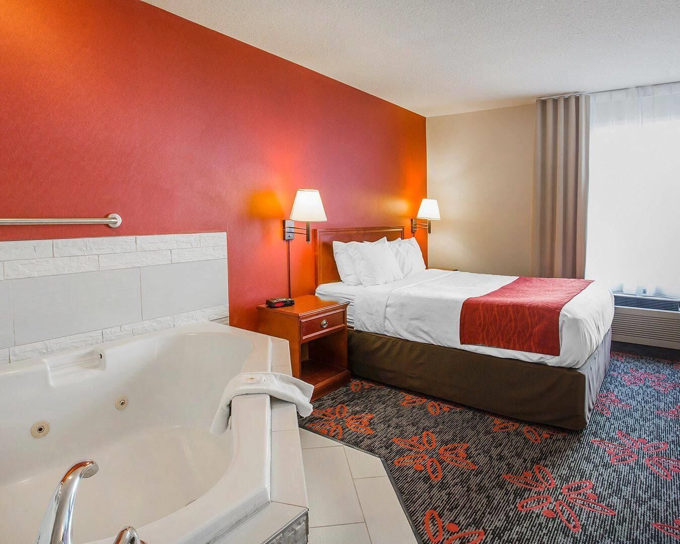A hotel room with a large bathtub, a bed with white linens, two bedside lamps, and a red carpet with a floral pattern.