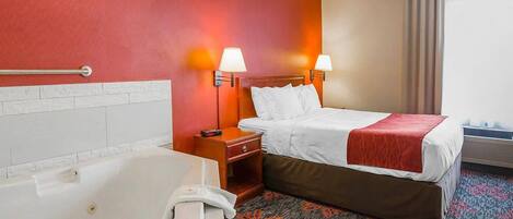 Habitación de hotel con una bañera grande, una cama con ropa blanca, dos lámparas de noche y una alfombra roja con motivos florales.