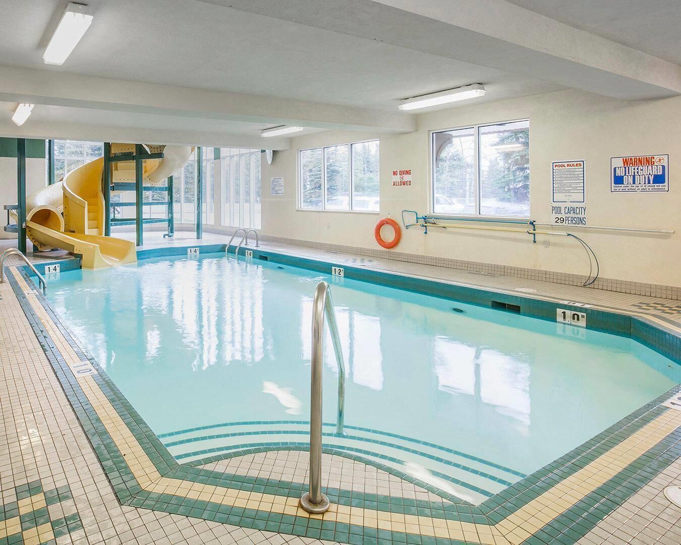 An indoor swimming pool with a slide, pool capacity sign, and lifeguard duty warning.