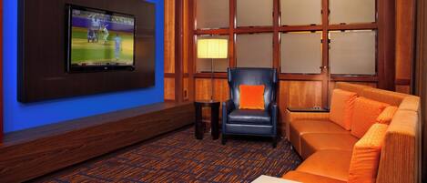 Habitación de hotel con una televisión de pantalla plana montada en la pared, un sillón de cuero, un sofá naranja y una alfombra estampada.