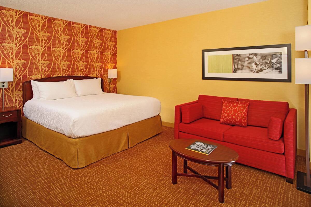 Una habitación de hotel con cama, sofá rojo, una mesita y un cuadro en la pared.