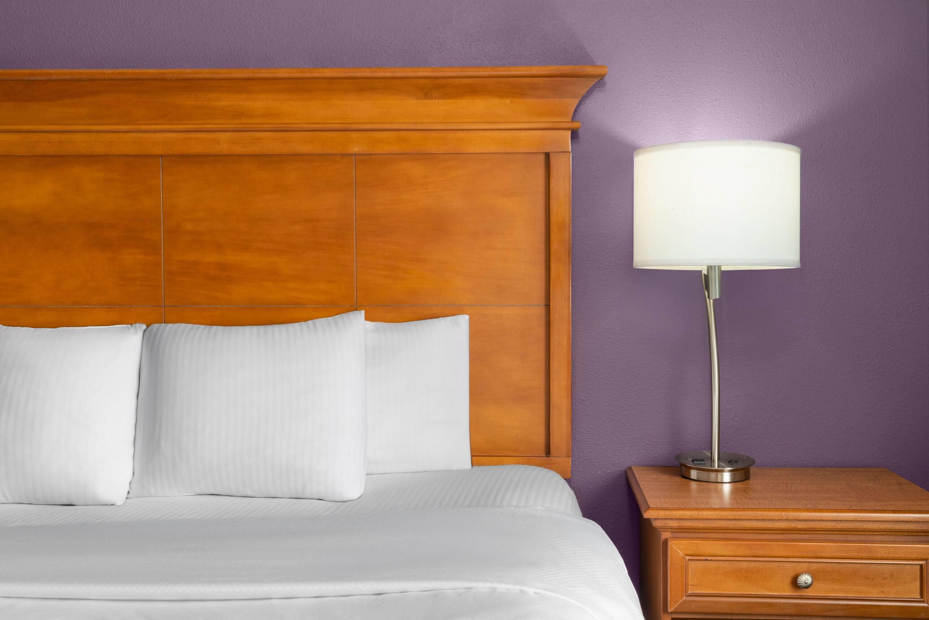 A neatly made bed with white linens and pillows, a wooden headboard, and a bedside lamp on a wooden nightstand.